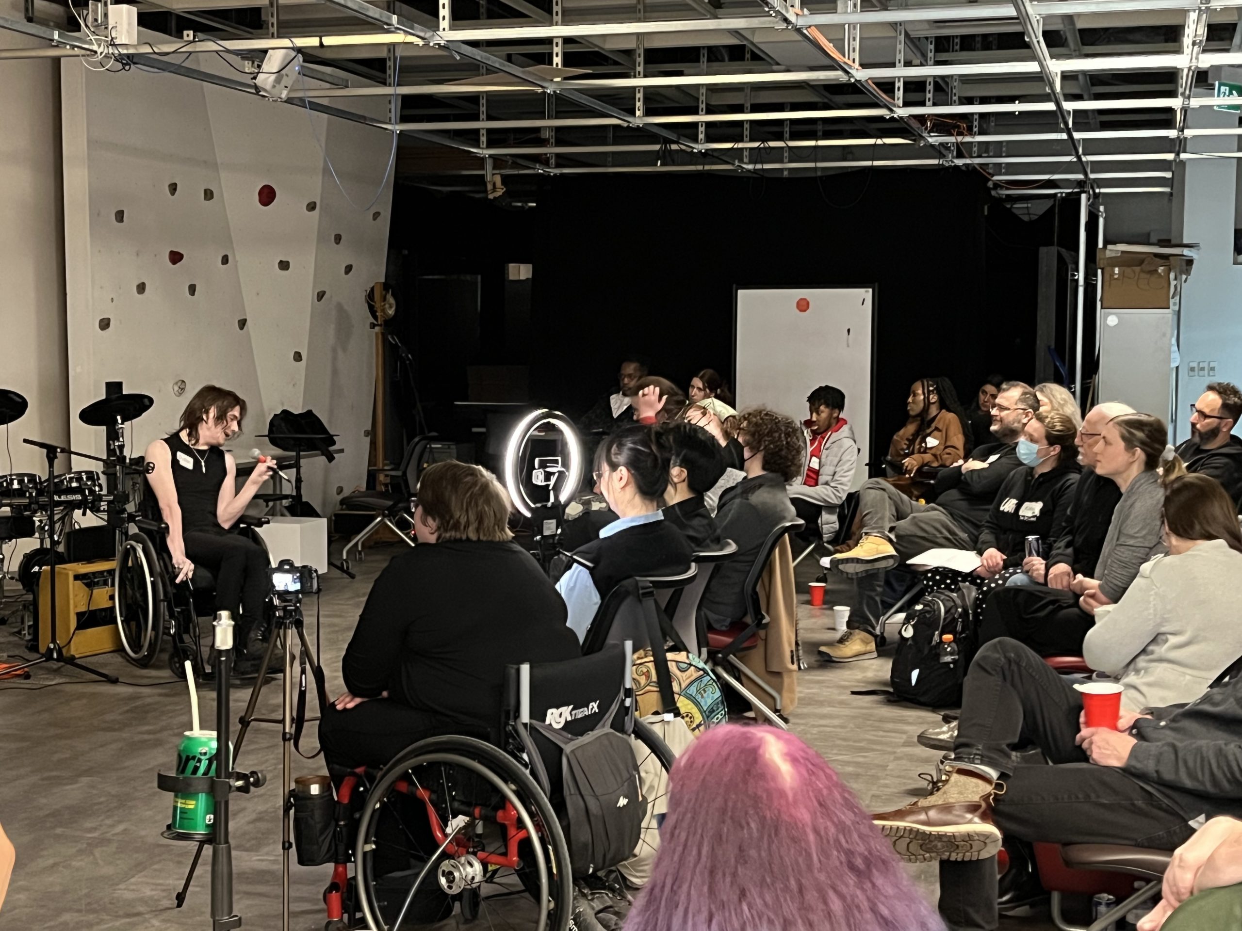 Dyllan Lambert Monroe sitting in Re/Lab, holding a microphone and giving a speech in front of a crowd of sitting people.
